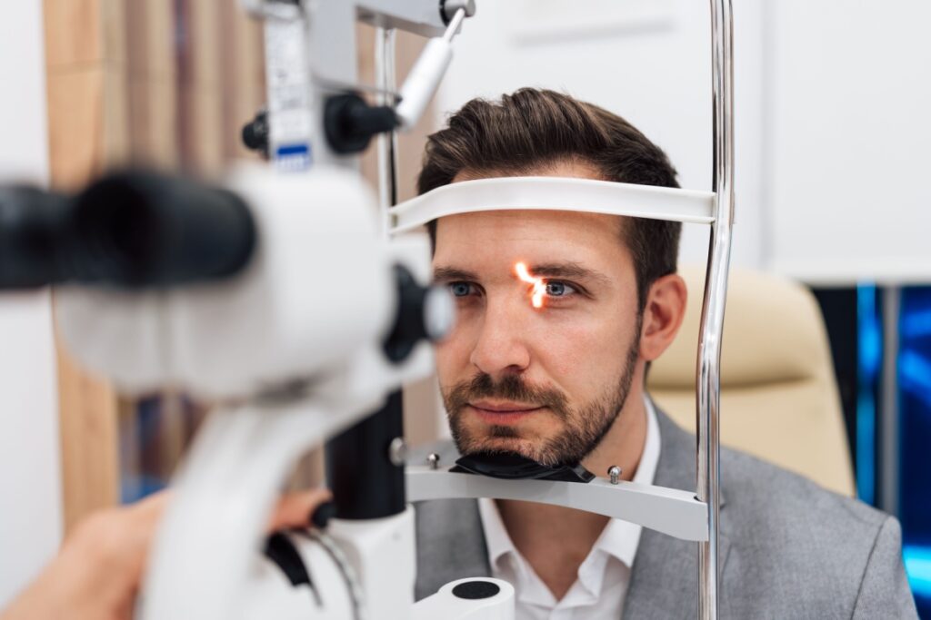 A patient sitting at a slit lamp while an optometrist examines the front of his eye with a focused light.