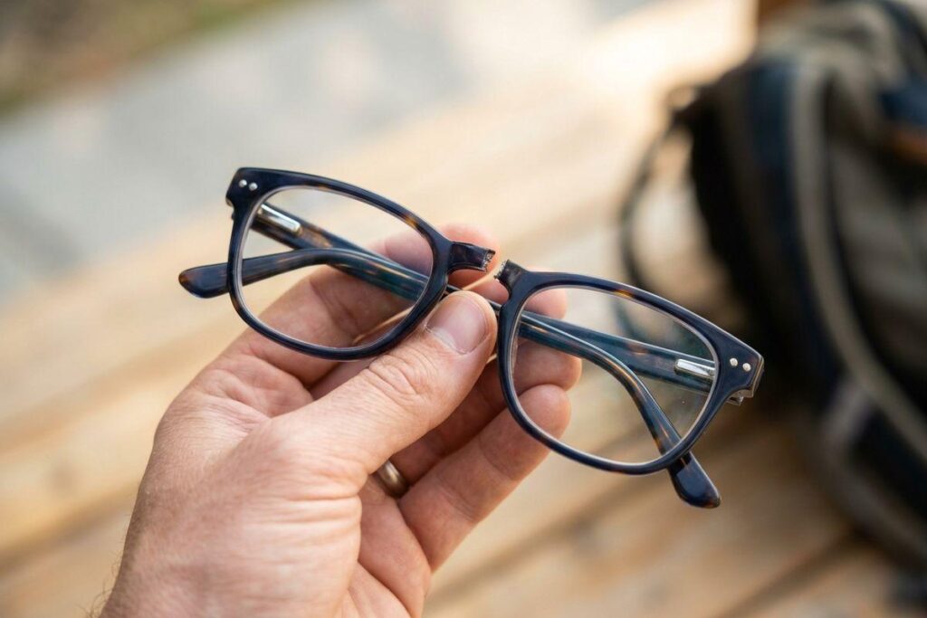 Hand holding a pair of eyeglasses broken at the bridge, with a blurred bag and wooden surface behind.